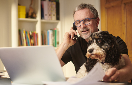 An older man works on his laptop while holding his dog.