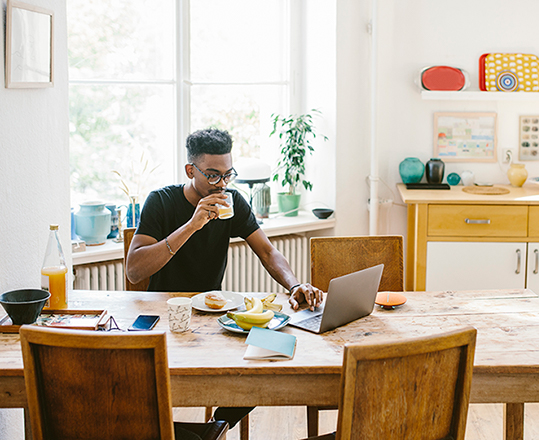 Man works at laptop on kitchen table while drinking juice