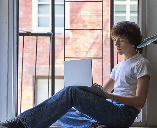 young man working on laptop and sitting on windowsill at home